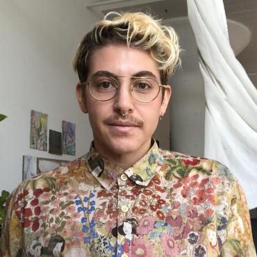 Sol Brager, a 34-year-old white, Jewish, trans cartoonist wearing a floral Henry Darger shirt and gold rimmed glasses, looks at the camera.