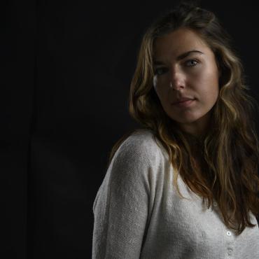 Kathryn Savage, a thirty-something white woman sits in front of a dark background, looking at the camera.