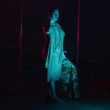A 30 year old Black transgender woman with a short geometric haircut stands in front of a mirror staring at herself in moody lighting. She is holding a dress up to her body and a jacket to her side.