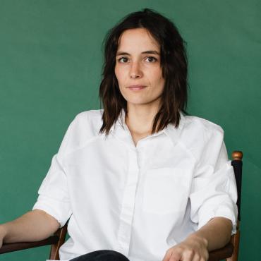 Simone LeClaire, a thirty-something white person with dark shoulder-length hair, dark eyes and wearing a white button-down looks at camera against a jade green background.