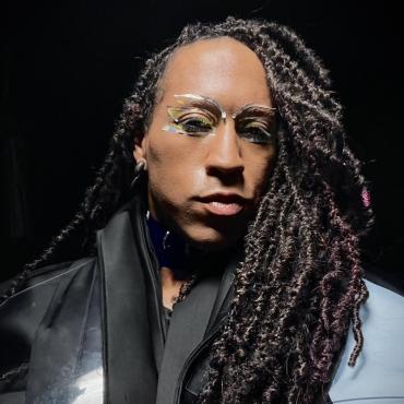 A mixed-race non-binary person looks directly into the camera. With asymmetrical hair in locs, cast to one side, framing a face featuring reflective make up.