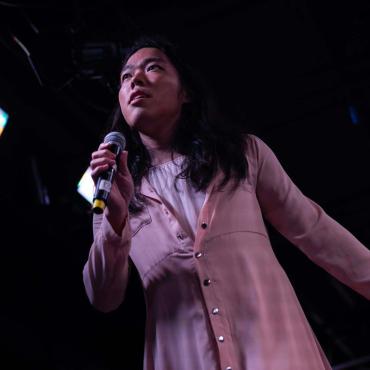 Softly holding a microphone in their hand, a young South East Asian trans woman gives a longing look above the horizon.