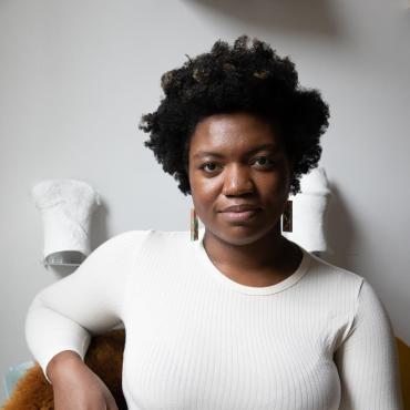 A dark skinned Black woman with short 4c hair is seated before a white wall with white plaster sculptures on it. She is wearing a white top and white pants and has a soft smirk on her face.
