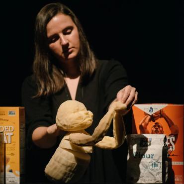 Yuliya Tsukerman, a White woman with brown hair wearing a black dress, puppeteers a bread baby made out of foam. The baby performs on raised black blocks surrounded by set pieces that include two cereal boxes and a bag of flower.