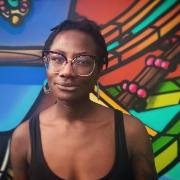 Raven Johnson, a thirty-something Black woman standing in front of a multi-colored wall.