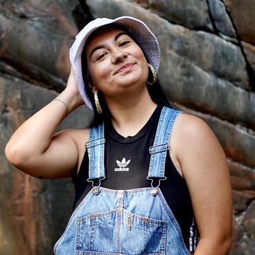 Sequoia is a 25-year-old queer, non-binary, trans, Native (Anishinaabe and Hupa) artist wearing overalls, a black Adidas tank top, a white bucket hat, gold septum and nose rings and sweetgrass hoops. Standing in front of a rock background looking into the camera with a slight head tilt and grin.