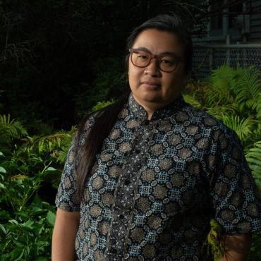 Valerie Oliveiro, 46, mixed-race non-binary Southeast Asian in a blue, black and gray batik shirt with a mandarin collar. Surrounded by summer ferns, the image is a late evening portrait. They have their hands in their pockets looking calmly at the camera.