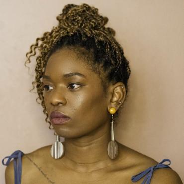 Charisse Pearlina Weston, a thirty-something Black women artist, sits in front of a mauve wall, in three quarter profile.