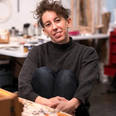 A white, genderqueer person smiling and holding their legs while seating in front of a weaving loom.