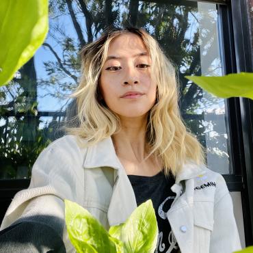 A selfie of Yo-Yo, a Taiwanese-American femme, sitting outside on her fire escape. It’s a sunny day, her hair is blonde and slightly wavy, she gazes serenely at the camera. Basil leaves frame the photo. Behind her is a window reflection of a tree and blue sky.