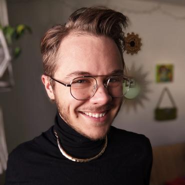 Merit, a 30-year-old white transsexual animator, smiles at the camera wearing a black turtleneck and gold necklace.