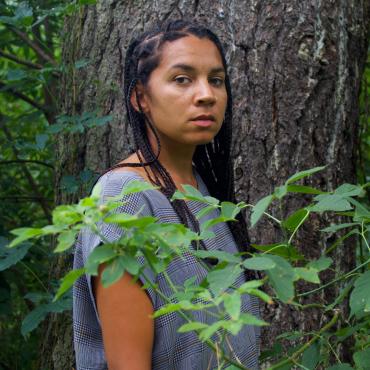 Lela Pierce, A Black multiracial woman in her mid 30's stands in front of a large tree trunk with some foliage in front of her.