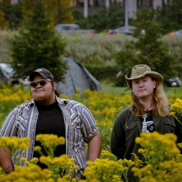 Jobi (Left), a twenty-something biracial trans man and Brandi (Right), a twenty-something white queer person stand in a field of golden rod while smirking at the camera.
