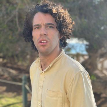 An outside headshot of Reid, a white man with curly hair. He stands outside in a yellow button-up shirt. The sun slightly flares into the shade of surrounding green trees.