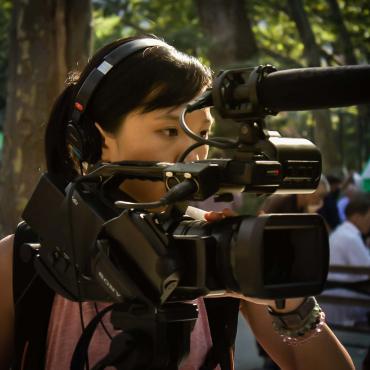 An Asian American woman filmmaker wearing headphones while operating a camera at an outdoor park in Manhattan Chinatown, New York City.