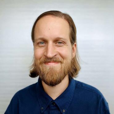 A headshot of Adam Loomis, a 34 year old white, male animator. He has chin-length brown hair tucked neatly behind his ears, a trimmed beard, and is wearing a navy blue button-up shirt.