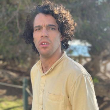 "An outside headshot of Reid, a white man with curly hair. He stands outside in a yellow button-up shirt. The sun slightly flares into the shade of surrounding green trees."