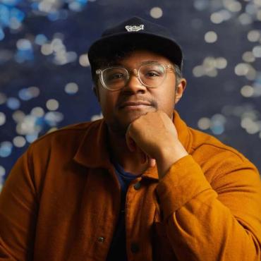 A headshot of Brit Fryer in a hat and glasses against a blue background.
