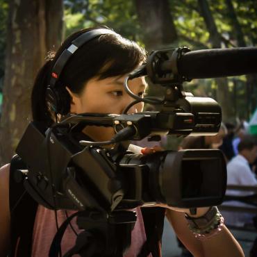 "An Asian American woman filmmaker wearing headphones while operating a camera at an outdoor park in Manhattan Chinatown, New York City."