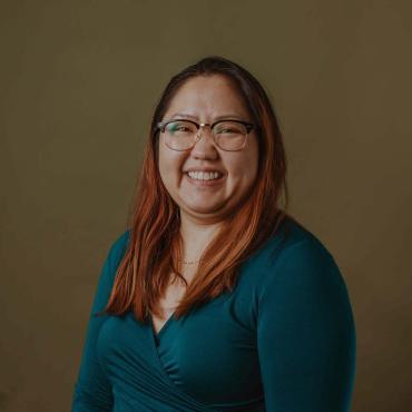 Joua, a 34 year old smiling Asian woman with blonde-orange hair with black roots, wearing glasses a bold teal shirt, in front of an olive background.