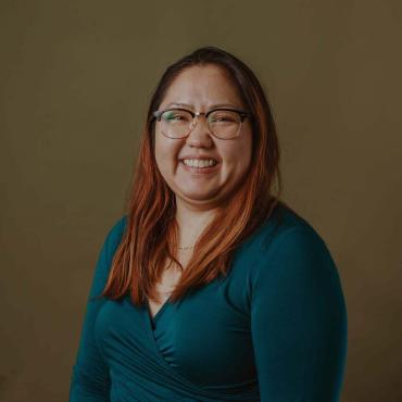 Joua, a 34 year old smiling Asian woman with blonde-orange hair with black roots, wearing glasses a bold teal shirt, in front of an olive background.