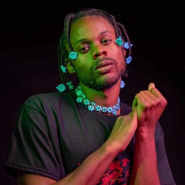 Prince Harvey stands wearing a black t-shirt with the image of a burning car on it. Masculine presenting, dark brown skin, a seductive look in the eyes. A shallow beard. Hair, box braided with blue flower barrettes and the tips. Hands clasped and held against chest.