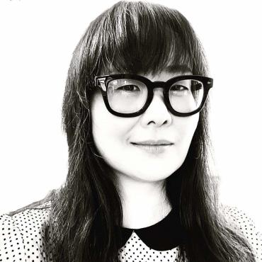 Headshot of Asian American woman with long hair and bangs, wearing black-rimmed glasses. She is wearing a black and white polka dot top.