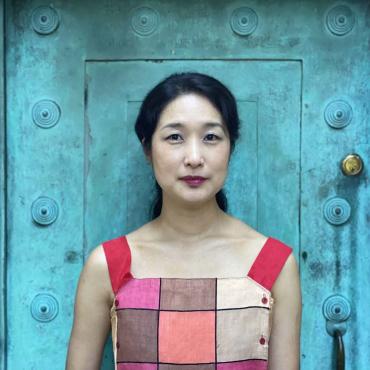 Headshot of artist Gamin, wearing sleeveless top-Korean modern hanbok, standing in front of blue door. The photo was taken at the Fort Greene park in Brooklyn.