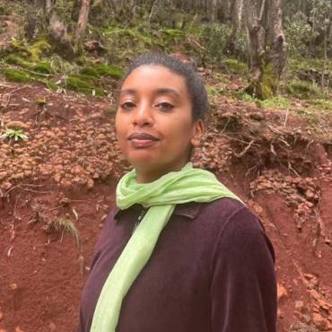 Hasabie, an image of the artist wearing a light green scarf and dark purple sweater. She is taking a walk in the forest with Eucalyptus trees in the background.