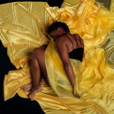 Leslie, a Black femme dance artist dressed in black attire and wrapped in yellow silk fabric with back exposed.