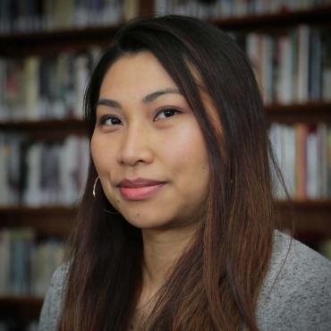 Headshot of the artist smiling in front of a bookshelf. She is wearing gold hoop earrings and a grey sweater.