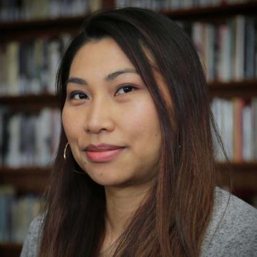 Headshot of the artist smiling in front of a bookshelf. She is wearing gold hoop earrings and a grey sweater.