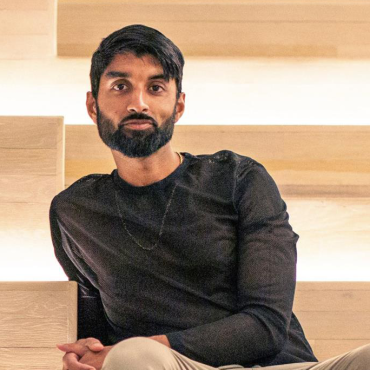 Anooj, South-Asian, bearded, and in his 30s, sitting on a set of steps wearing tan pants and a black shirt, looking at the camera.