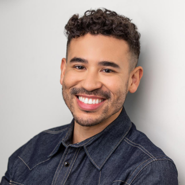 A friendly close-up of a man with short, curly dark hair and a well-groomed beard. He has warm brown eyes and a subtle smile, giving off a relaxed and approachable vibe. He’s wearing a dark denim button-up shirt with light stitching.