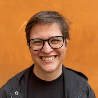 Andrea is wearing glasses and all black, smiling against an orange wall.