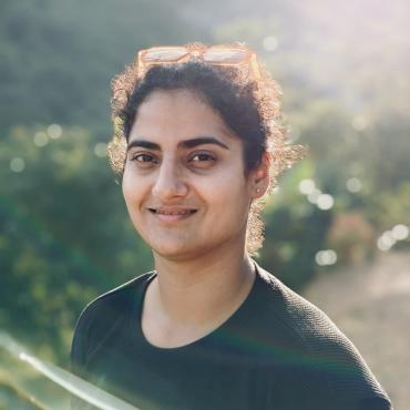 A South Asian woman with curly hair pulled back smiles while standing outdoors in natural light. She is wearing a dark shirt with tan sunglasses resting on her head, framed by a lens flare and blurred greenery in the background.