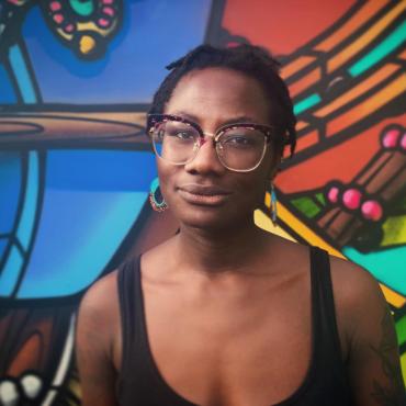 Raven Johnson, a thirty-something Black woman with glasses, stands in front of a colorful mural.