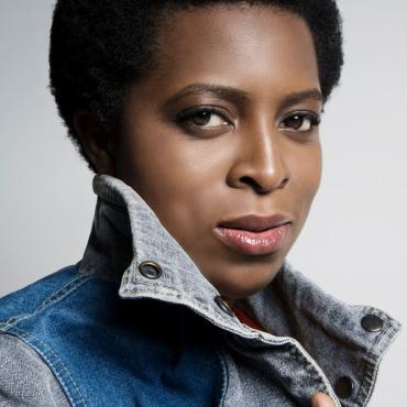 A Black woman looks toward camera in a jean vest smiling.