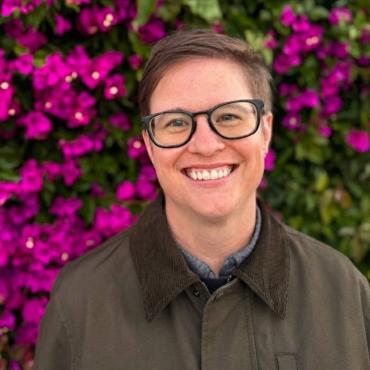 Andrea is wearing glasses and smiling, with a wall of flowers and leaves behind her.