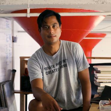 In the foreground, Witt Siasoco is staring into the camera wearing a grey Private Property Created Crime t-shirt. In the background is a brightly colored studio with sketches and paintings hung and piles of books stacked on a desk.