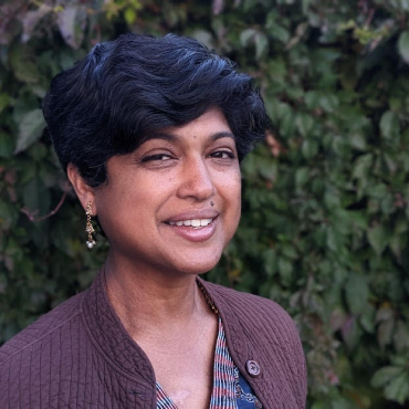 Pramila Vasudevan smiling, standing against a wall of greenery