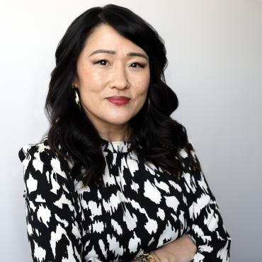 Photograph of an Asian femme person with longish hair, wearing a white and black shirt, standing with arms crossed